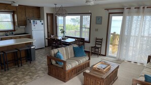 Uma sala de estar ampla com sofá de madeira, mesa de centro e mesa de jantar. Cozinha com armários de madeira e uma geladeira é visível. Janelas grandes oferecem vista para o exterior.
