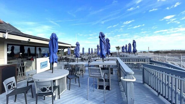 Een terras buiten met tafels en stoelen, blauwe parasols en een hek met uitzicht op een waterpartij.