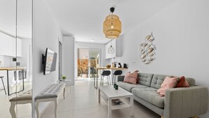 Een moderne woonkamer met een grijze bank, een witte salontafel en een zitbank. Er is een eethoek met een tafel en stoelen zichtbaar door een open deur.