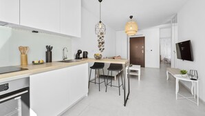 Een moderne keuken met witte kasten, een houten aanrechtblad en zwarte barkrukken. Er is een eethoek met een witte tafel en een bank, en een tv die aan de muur hangt.