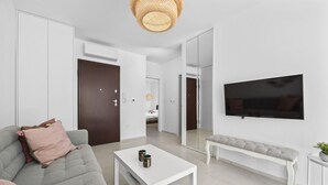 Een moderne woonkamer met een grijze bank, een witte salontafel en een bankje. Er is een flatscreen televisie aan de muur en een grote spiegel die de kamer weerspiegelt.