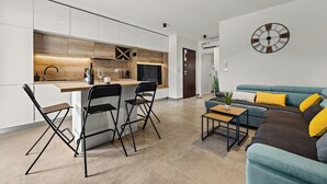 A modern kitchen with white cabinets, a wooden countertop, and black bar stools. There is a blue sofa with yellow cushions in the living area, and a large wall clock. - Couple's Vacation with Jacuzzi - Crepsa Paradise (Cres)