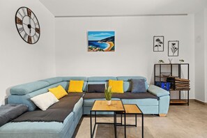 A modern living room with a blue sectional sofa, a wooden coffee table, a wall clock, and a painting of a coastal landscape. - Couple's Vacation with Jacuzzi - Crepsa Paradise (Cres)