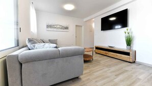 Uma sala de estar moderna com um sofá cinzento, um móvel de televisão em madeira e uma janela grande.
