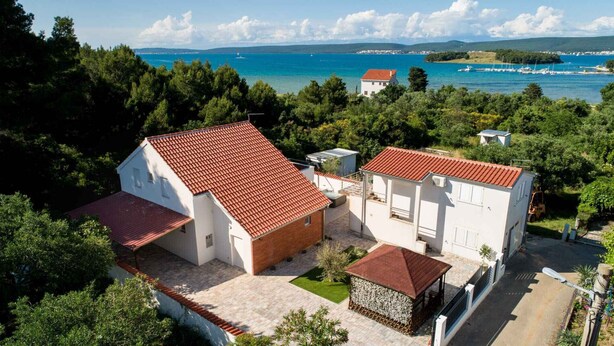 Uma casa de dois andares com telhado de telhas vermelhas, cercada por vegetação e vista para uma área costeira com barcos.
