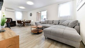 Uma sala de estar moderna com um sofá seccional cinza, uma mesa de centro de madeira, uma mesa de jantar com cadeiras e uma grande janela com cortinas leves.