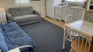 A compact living space with a sofa, a bed, a kitchenette with a fridge, stove, and sink, and a window with blinds. - Cozy cabin less than 100 steps to the sand! (Hampton)
