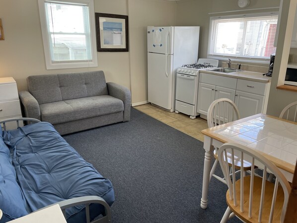 A compact living space with a sofa, a bed, a kitchenette with a fridge, stove, and sink, and a window with blinds. - Cozy cabin less than 100 steps to the sand! (Hampton)