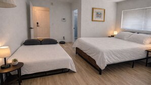 Une chambre avec deux lits, une table de chevet avec une lampe, une petite plante et une porte donnant sur une autre pièce.