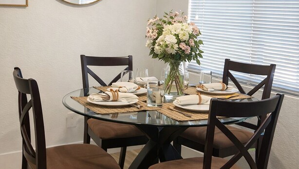 Un coin repas comprenant une table ronde en verre, des chaises en bois foncé, un ventilateur de plafond, un miroir rond et une fenêtre avec des stores.