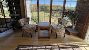 Une pièce dotée d’un plancher en bois, de deux fauteuils à motifs, d’une petite table et offrant une vue sur un jardin grâce à de grandes fenêtres.