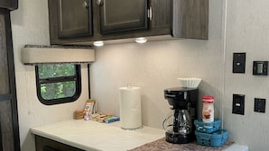 A compact kitchenette with dark wooden cabinets, a countertop with a paper towel holder, a coffee maker, and a small refrigerator. - Farm Glamping @ The Sage Getaway RV (Hot Springs)
