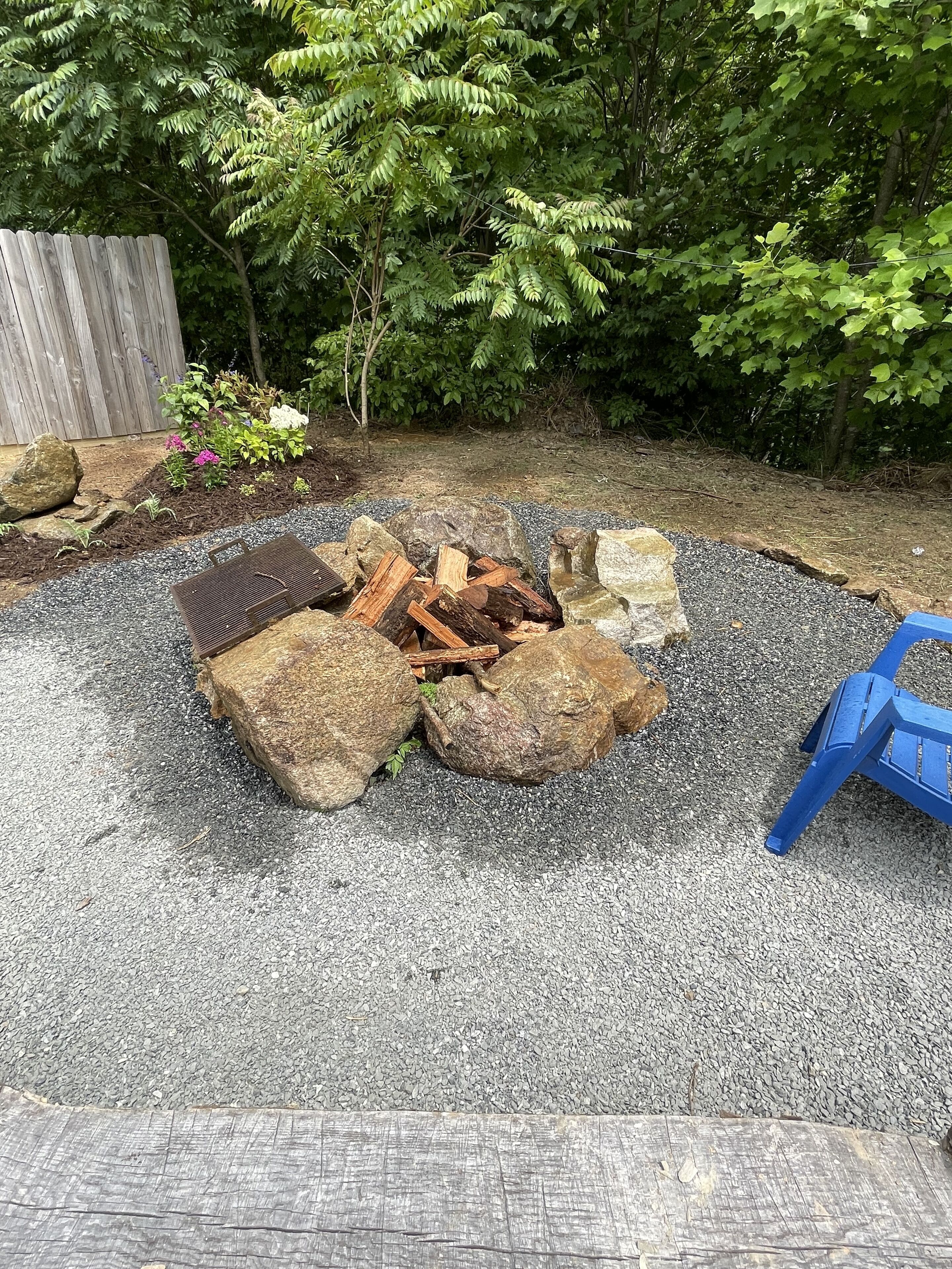 A garden with a fire pit surrounded by rocks and logs, a wooden fence, and various plants.