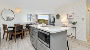 A modern kitchen with a built-in microwave, a dining area with wooden chairs, and a large window offering a view. - Aloha Chic Penthouse Vistas (Kihei)