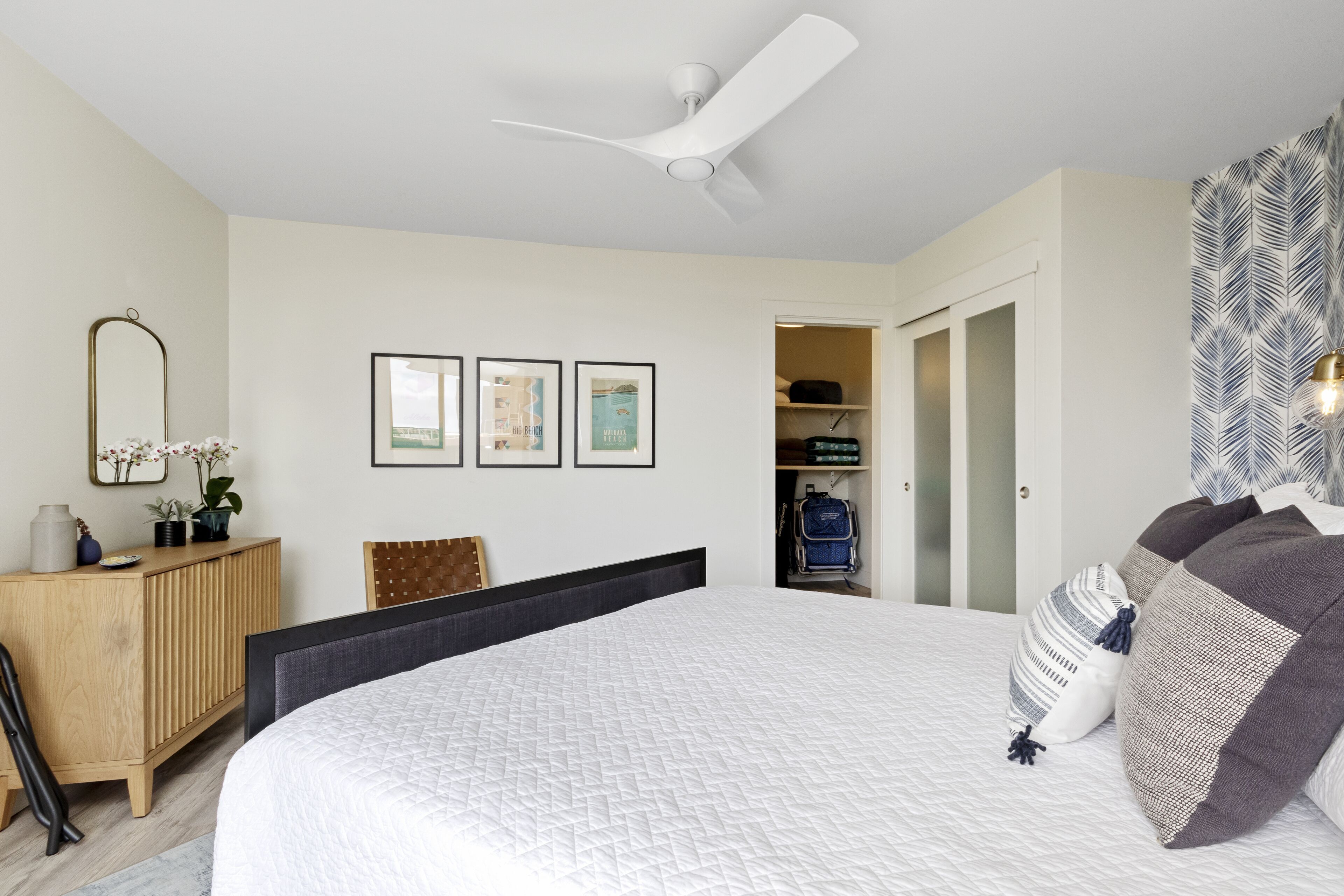 A bedroom with a large bed, a ceiling fan, a wardrobe, and a desk with a mirror and plants.