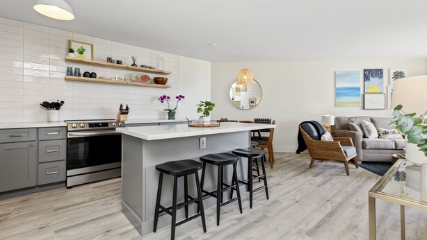 A modern kitchen with a central island, bar stools, a dining area with a round table, and a living room with a sofa and decorative items. - Aloha Chic Penthouse Vistas (Kihei)