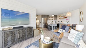 A modern living room with a large TV, a dining area, and a kitchen. - Aloha Chic Penthouse Vistas (Kihei)