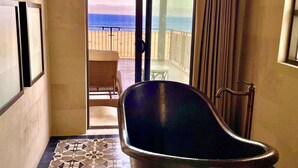 Une salle de bain avec une baignoire, un carrelage décoratif et une vue sur la plage, visible à travers une porte vitrée.