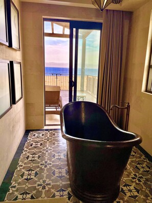 Een badkamer met een bad, een vloer met een patroon, en uitzicht op het strand door een glazen deur.