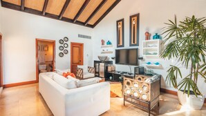 A living room with a white sofa, a TV, and a wooden coffee table. There is a door leading to a bathroom, and a large plant is visible. - Penthouse w Jacuzzi and Ocean View Puerto Bahia (Samaná)