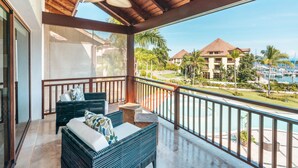 A balcony with wicker furniture, a ceiling fan, and a view of a resort with palm trees and a building in the background. - Penthouse w Jacuzzi and Ocean View Puerto Bahia (Samaná)