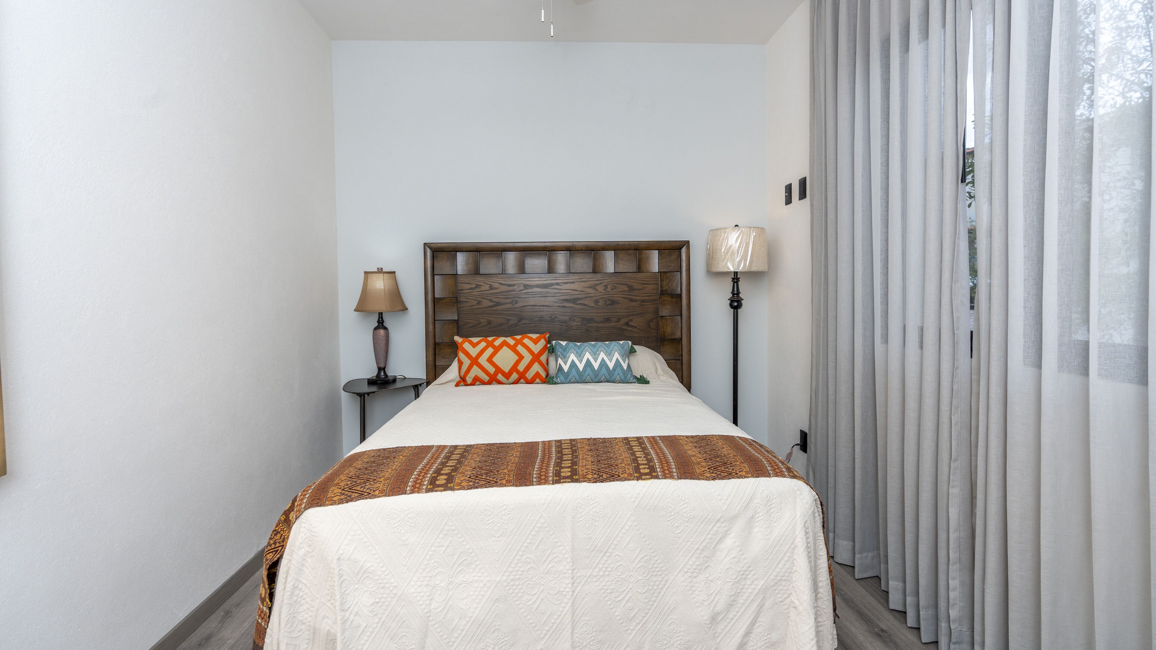 A neatly arranged hotel room with a wooden headboard, two bedside lamps, a patterned bedspread, and a view through sheer curtains.