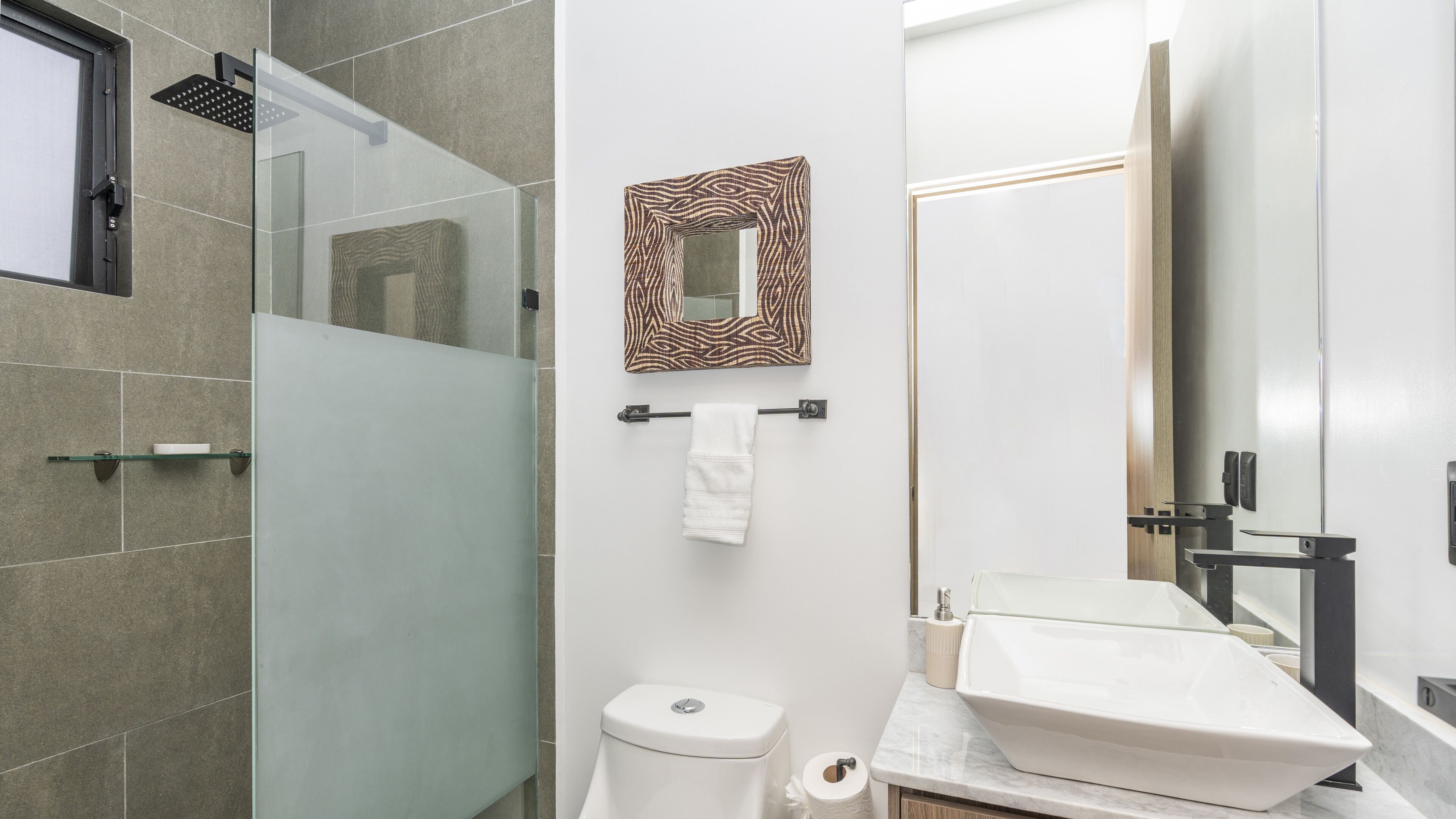 Un baño moderno con cabina de ducha de vidrio, inodoro, lavamanos con espejo grande y toallero.