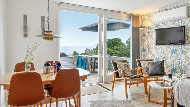 Ein modernes Wohnzimmer mit Essbereich, große Schiebeglasfenster, die auf einen Balkon mit Meerblick führen, ein an der Wand befestigter Flachbildfernseher und verschiedene Sitzmöglichkeiten.