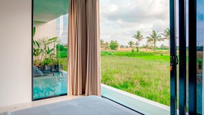 A room with a large glass door leading to a balcony with a view of a green field and palm trees. - Green Flow Villa 3 (Ubud)