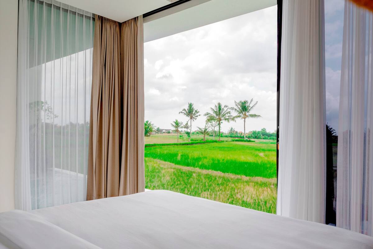 A bedroom with a large window offering a view of a green field and palm trees.