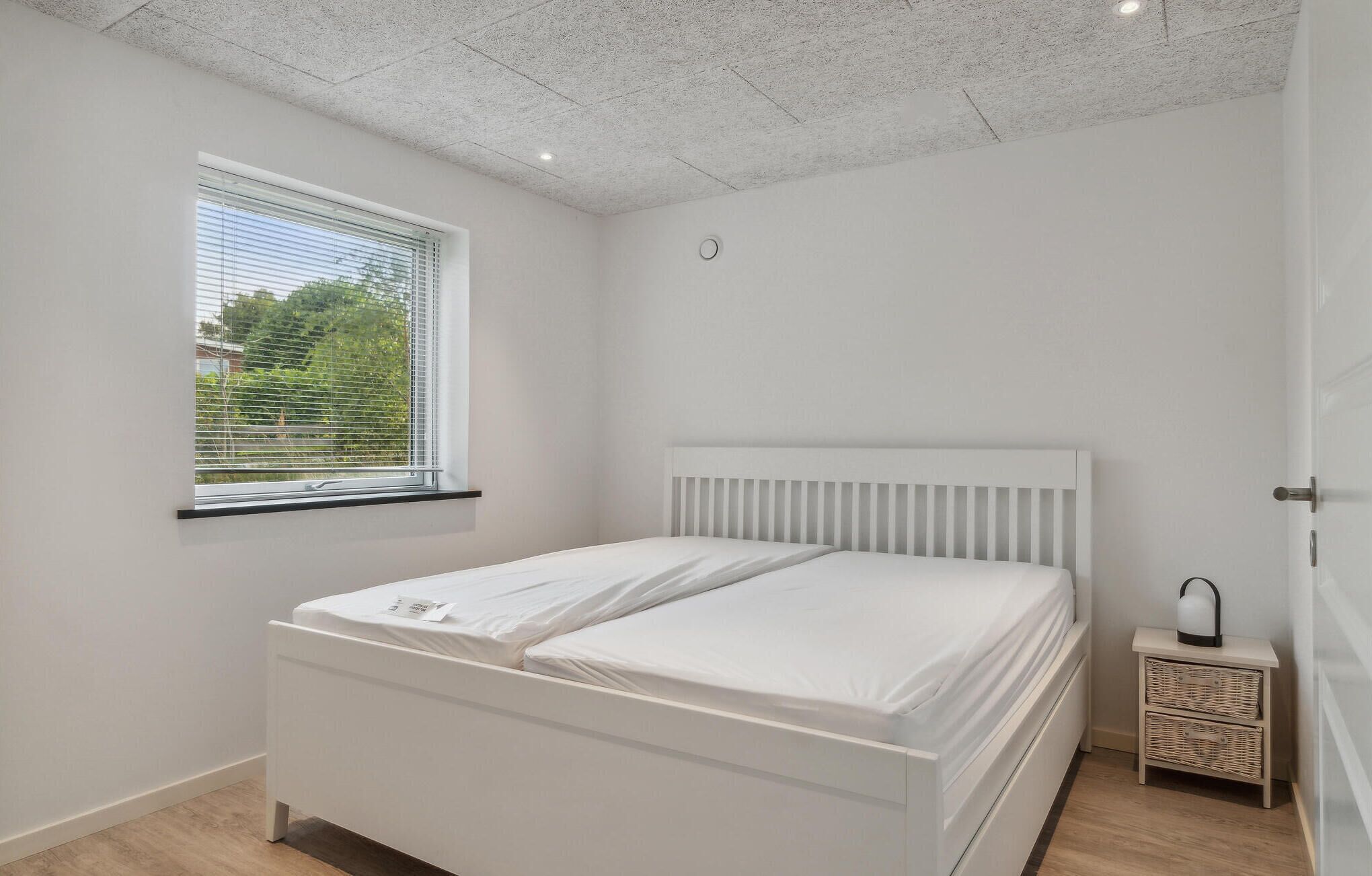 Quarto com cama branca, janela com persianas, mesa de cabeceira pequena com abajur e iluminação embutida no teto.