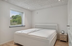 Ein Schlafzimmer mit einem weißen Bett, einem Fenster mit Jalousien, einem kleinen Nachttisch mit Lampe und einer Decke mit Einbauleuchten.
