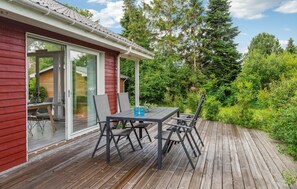 Eine Holzterrasse mit Tisch und Stühlen, eine Glastür, die zu einem überdachten Bereich führt, und ein üppiger Grünpflanzen-Garten im Hintergrund.