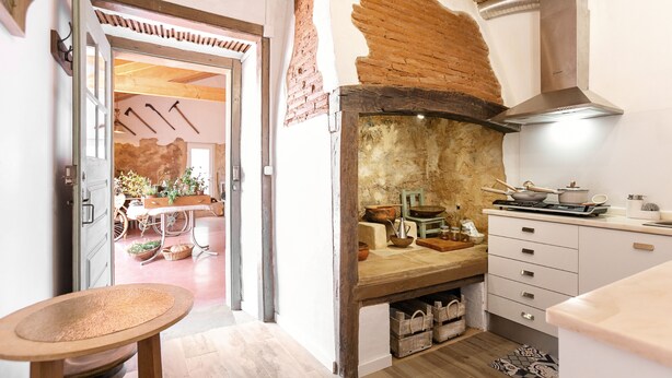 Une cuisine avec un mur en briques rustiques, des poutres en bois et un poêle moderne. On aperçoit une table en bois et une chaise au premier plan.