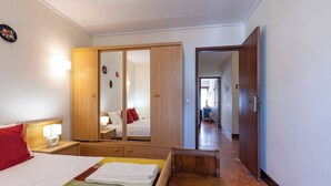 A hotel room with a bed, a wooden wardrobe, a bedside table with a lamp, and a view into the hallway. - Lighthouse Apartment (Sesimbra)