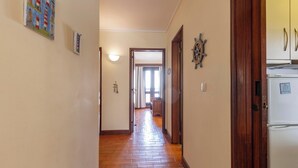 A hallway with wooden flooring, a white refrigerator, and a door leading to a room with a window. - Lighthouse Apartment (Sesimbra)