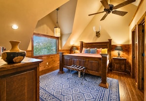 Una camera da letto con soffitto spiovente, un letto, una cassettiera in legno, un vaso, una finestra con persiane e un tappeto decorato.