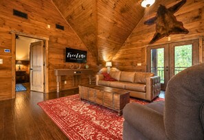 Interno di una baita di legno con un divano in pelle, un tappeto rosso decorato, una televisione a parete e una grande pelle di animale a soffitto.