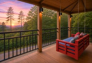 A wooden deck with a red swing bed and a view of a sunset over a forest.