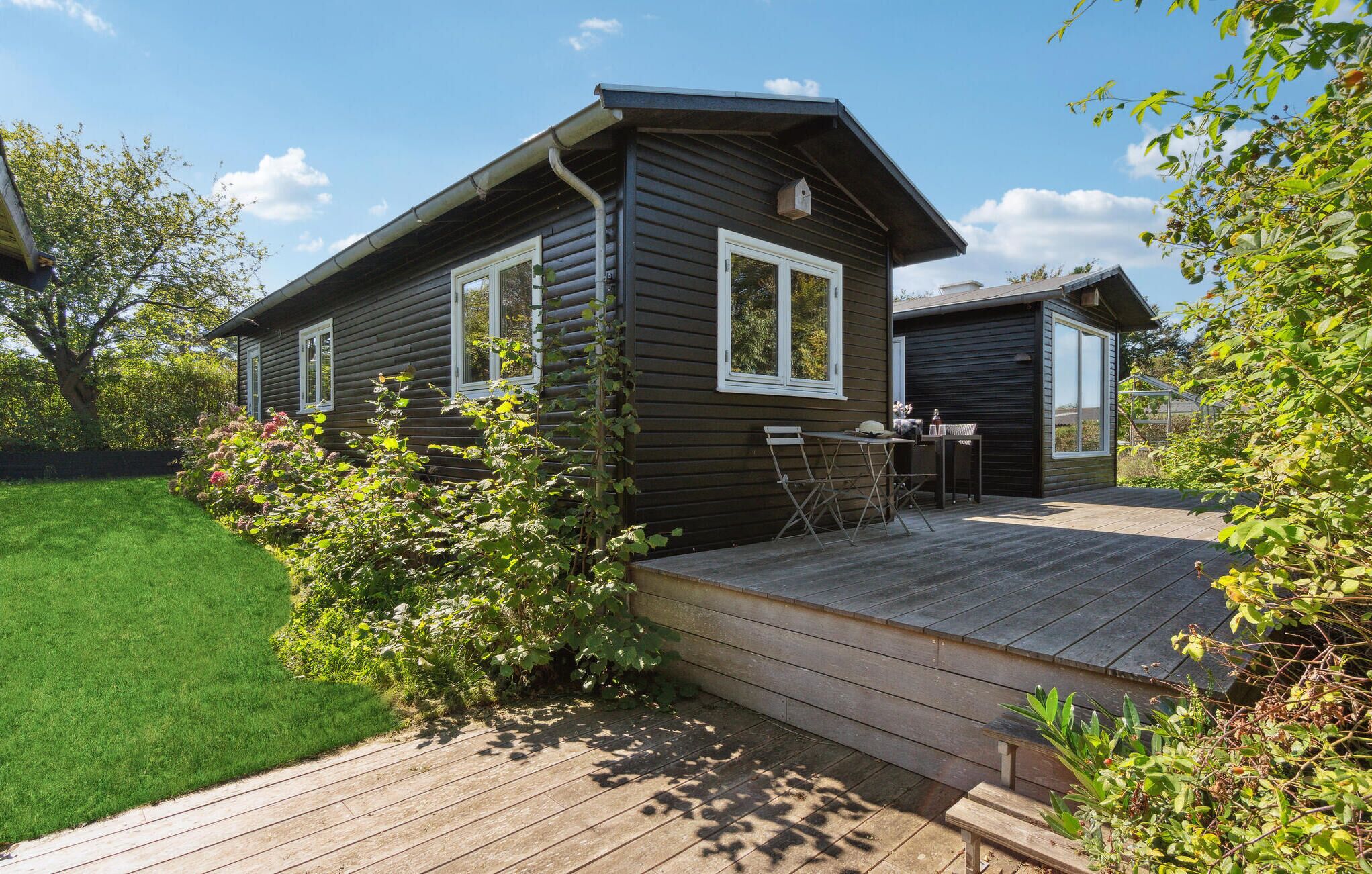 A wooden cabin with a deck, surrounded by greenery and a clear sky.