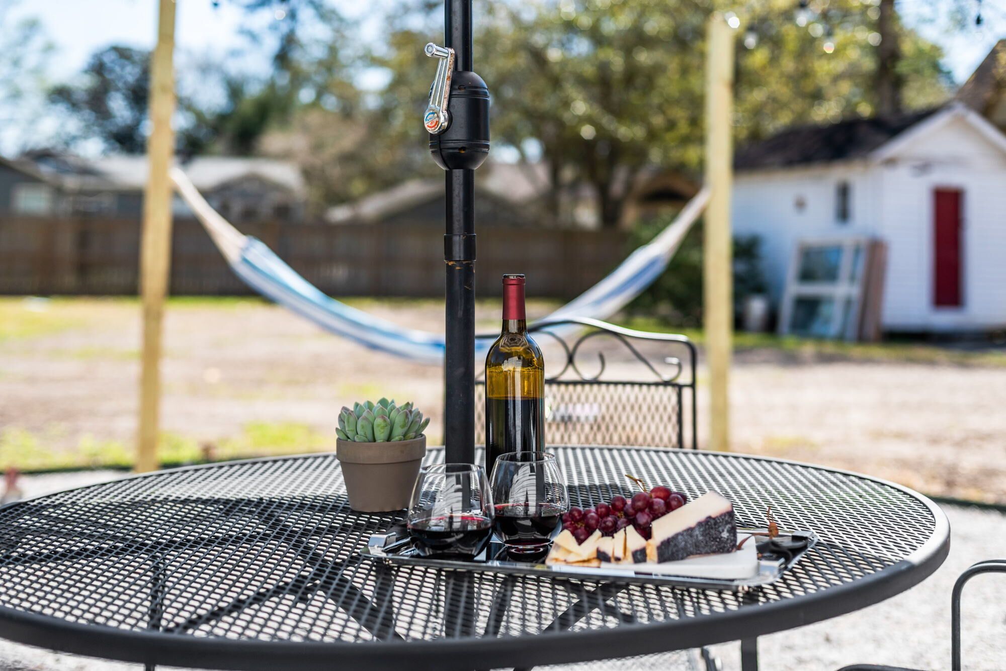 Une table ronde en métal, avec une bouteille de vin, des verres, une petite plante en pot et un morceau de fromage accompagné de raisins.