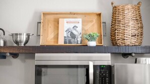 Un comptoir de cuisine avec une bouilloire en acier inoxydable, une passoire, un plateau en bois sur lequel repose un livre, une petite plante en pot et un panier tressé.