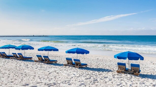 Ein Strand mit Reihen von Liegestühlen und blauen Sonnenschirmen, einem klaren Himmel und ruhigen Wellen.