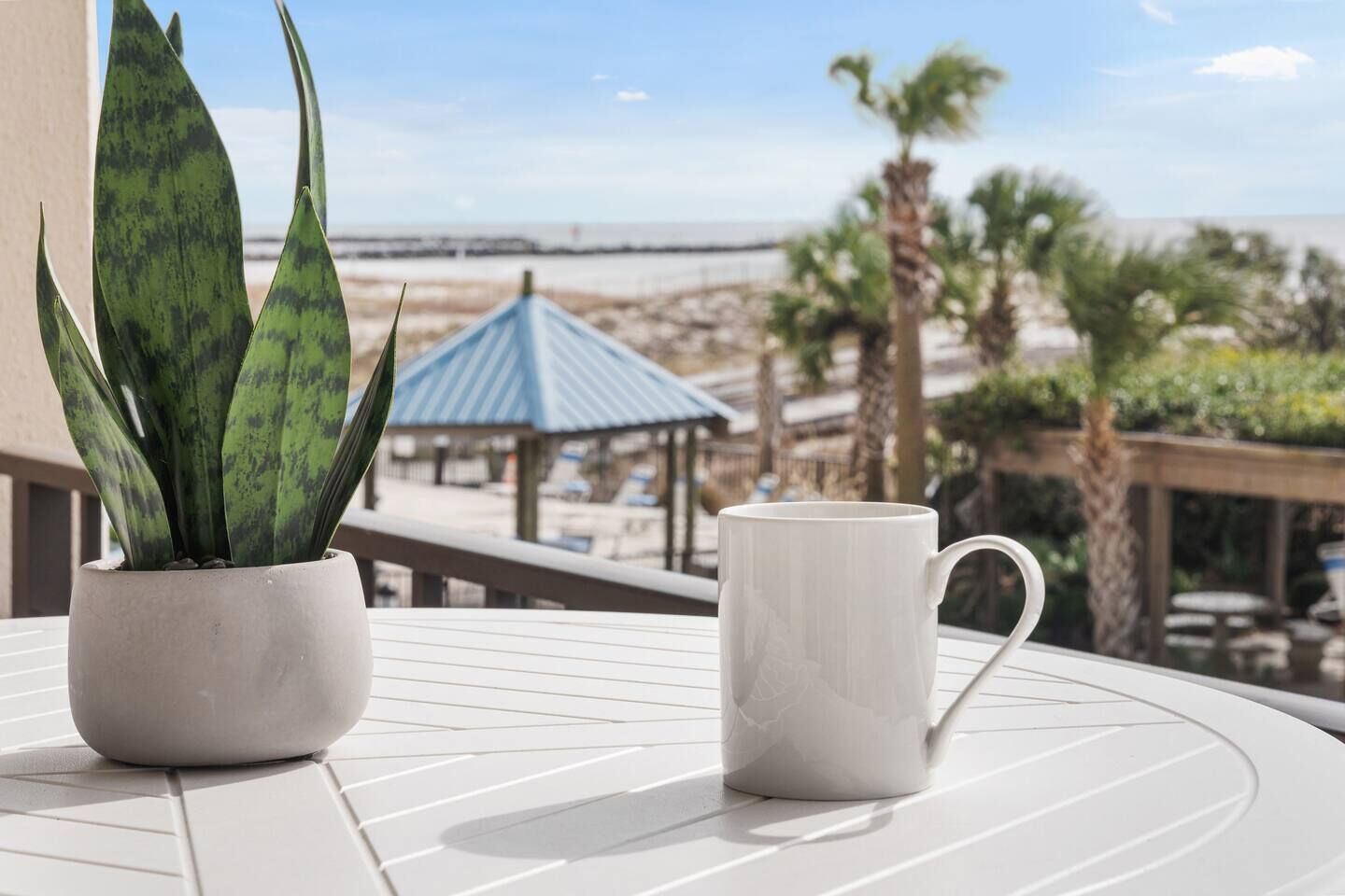 A potted plant and a mug on a white table with a view of a beach and a gazebo.