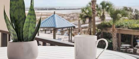 A potted plant and a mug on a white table with a view of a beach and a gazebo.