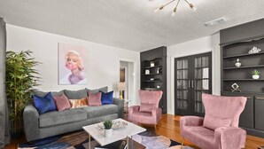 A modern living room with a grey sofa, two pink armchairs, a coffee table, and a bookshelf. - 1 Acre Estate Across the Lake from LSU (Baton Rouge)