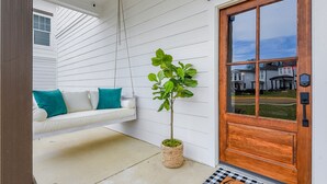 Un porche à l’extérieur blanc, une porte en bois, une plante suspendue et un canapé blanc agrémenté de coussins turquoise.