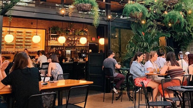 Uma área de refeições ao ar livre com mesas e cadeiras, um bar com prateleiras cheias de garrafas e plantas penduradas.