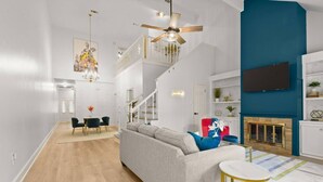 Uma sala de estar moderna com um sofá cinzento, uma área de jantar com cadeiras, uma escada, uma TV na parede e uma lareira.