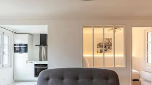 A modern kitchen with white cabinetry and stainless steel appliances, a dining area with a grey upholstered chair, and a window with a cityscape view. - La petite source: modern apartment in the heart of Honfleur (Honfleur)